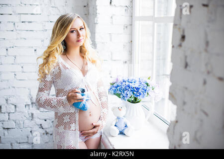 Charmante schwangere Mädchen steht am Fenster mit Kinderschuhe. Stockfoto