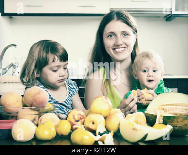 Glückliche Mutter mit Kindern Essen Melonen und anderen Früchten über Tisch zu Hause Stockfoto