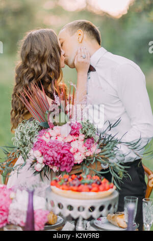 Die Nahaufnahme Porträt des Küssens Brautjungfer und der beste Mann hinter den Hochzeitstisch. Die BRAUTJUNGFER das Bouquet von päonien. Frühjahr Zusammensetzung Stockfoto