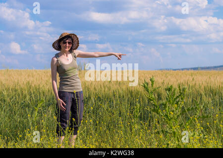 Kaukasische Frau mit Hut zeigen mit dem Finger auf den blauen Himmel in ein Feld. Stockfoto