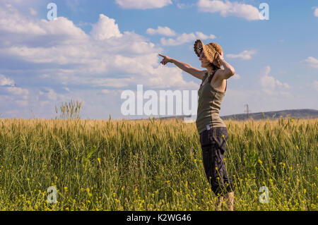 Kaukasische Frau mit Hut zeigen mit dem Finger auf den blauen Himmel in ein Feld. Stockfoto