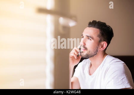 Stattliche latino im weißen T-Shirt, am Telefon zu sprechen. Junge spanische Mann mit Bart sitzt in der Sonne durch offene Fenster im Schlafzimmer und Außerhalb suchen. Stockfoto