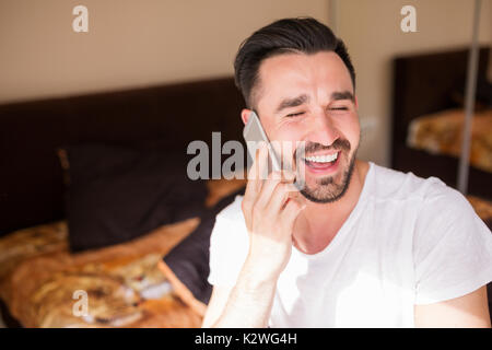 Attraktive Bachelor während Anruf lachen. Seine Augen sind geschlossen und den Mund offen in großen toothy Lächeln. Er trägt weiße t-shirt als Schlafanzug. Es gibt Stockfoto