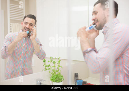 Attarctive latin Mann per Telefon Gespräch während die Zähne putzen. Schönes häusliches Bad ist hell erleuchtet. Der Mensch ist in die Spiegel Reflexion sichtbar. Stockfoto