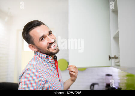 Lächelnd Mann in Küche durch den Schrank. Weiße, moderne Küche mit glänzenden Holzmöbeln und weißen Steinen an die Wand. Stockfoto