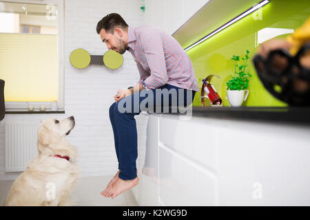 Mensch und Hund einander in der Küche. Mann sitzt auf einer Arbeitsplatte in der Küche und ein Hund sitzt vor einem Mann. Stockfoto