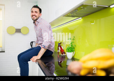 Hübscher junger Mann sitzt auf einer Arbeitsplatte in der Küche und lächelte. Er ist legere kariertem Hemd und blauer Hose. Er ist bärtige Latino. Er ist in seinem Stockfoto