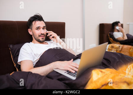 Lächelnd Mann im Bett liegend mit Laptop und am Telefon zu sprechen Stockfoto