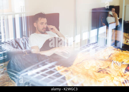 Mann im Bett liegend mit Laptop und am Telefon zu sprechen Stockfoto