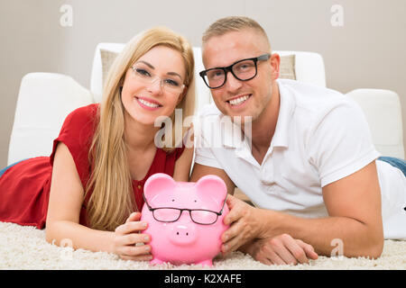 Portrait von glücklichen Paar mit Sparschwein auf dem Teppich liegend Stockfoto