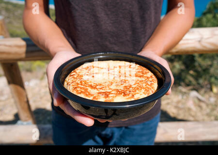 Nahaufnahme eines jungen kaukasischen Mann im Freien mit einer Platte mit einem typischen Tortilla de patatas, spanisches Omelette, in seinen Händen, mit dem Meer in der backgroun Stockfoto