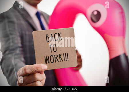 Nahaufnahme eines jungen kaukasischen Geschäftsmann trägt einen Schwimmring in der Form eines rosa Flamingo mit einem braunen Schild mit dem Text zurück zum vollstreckungsbescheid arbeiten Stockfoto