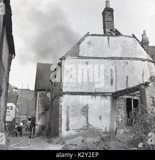 1964, historische, Bild zeigt einen Feuerwehrmann in einen Durchgang mit einem Schlauch zu Besprühen eines alten alten Elisabethanischen Fachwerk Gebäude mit Rauch aus dem Dach, Silver Street, Aylesbury, Herts, England, UK. Stockfoto