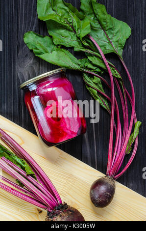 Hausgemachte Konserven rote Bete in klaren Gläsern mit frischen BIO Rote Bete auf schwarzem Holz- Hintergrund der Ansicht von oben. Stockfoto