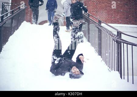 Ein Student sitzt im Schnee mit Beinen in der Luft nach unten schieben die verschneite Treppe, während andere Studenten im Schnee Gang stehen an der Spitze der Treppe gekleidet, auf einem Snow Day an der Johns Hopkins University, Baltimore, Maryland, 2016. Stockfoto