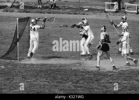 Graustufen Aktion Foto während einen Men's lacrosse Übereinstimmung zwischen der Johns Hopkins University und der United States Military Academy in West Point, als Micky Webster Nummer 10 von JHU schleudert die Kugel aus seinem Stock einen Schuß zu nehmen, während Armee Verteidiger und Torwart ihn und versuchen, den Schuss zu blockieren, mit anderen Spielern, darunter John jory Nummer 21 von JHU, folgenden eng hinter, Baltimore, Maryland, 27. April 1957. Stockfoto