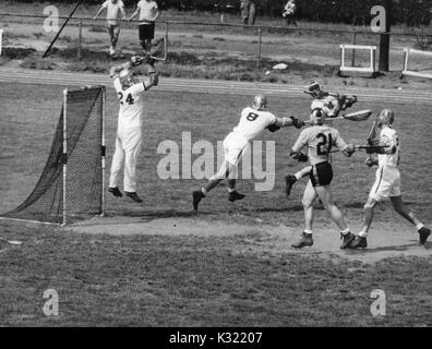 Graustufen Aktion Foto während einen Men's lacrosse Übereinstimmung zwischen der Johns Hopkins University und der United States Military Academy in West Point, während Micky Webster Nummer 10 von JHU schießt den Ball über seine Schulter, wodurch goalie und Verteidigung Mitglied aus der Armee in die Luft zu springen, um den Schuß zu blockieren, mit John jory Nummer 21 von JHU läuft das Ziel, Baltimore, Maryland, 27. April 1957. Stockfoto