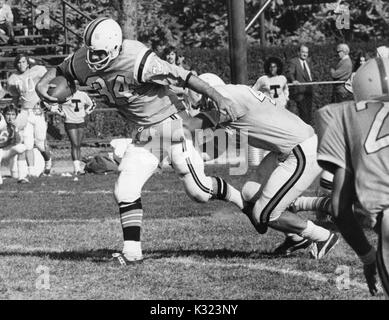 Ein Johns Hopkins University Football Spieler gegen einen Spieler des gegnerischen Teams mit dem Fußball mit Spieler und Fans an der Seitenlinie im Hintergrund an der Johns Hopkins University, Baltimore, Maryland, 1980 sichtbar. Stockfoto