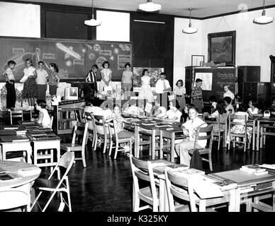 An eine elementare Demonstration school der Johns Hopkins Universität, Studenten in Herr Fuller's Strawbridge sechsten Klasse Raum Akademie sitzen am Schreibtisch, während eine Linie von Studenten präsentiert ihre Arbeiten an der Vorderseite des Klassenzimmers, mit einer Tafel mit Platz Abbildungen hinter Ihnen, und Herr Strawbridge am Ende der Leitung die Leitung der Aktivitäten, Baltimore, Maryland, Juni, 1955. Stockfoto