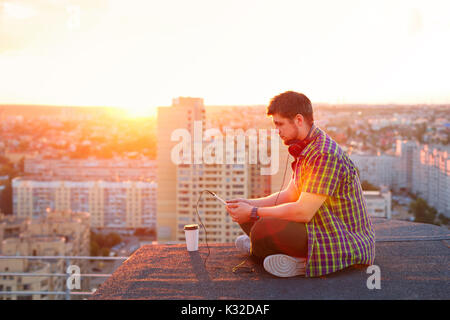 Hipster Mann Musik hören mit Kopfhörern auf dem Dach bei Sonnenuntergang. Er hält einen Tablet-PC in der Hand und liest die Nachrichten. Stockfoto