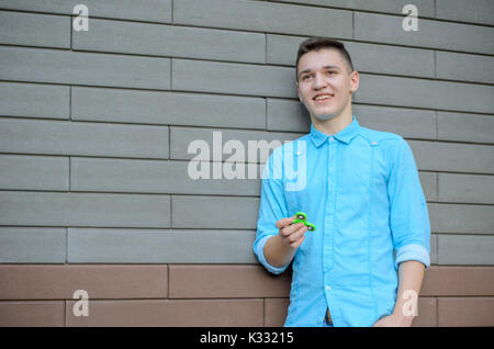 Lachend Teenager auf der Azure T-Shirt und Blue Jeans Hand beliebte Gadget zappeln Spinner. Mann spielt mit grünen Spinner im Freien auf der bric Stockfoto