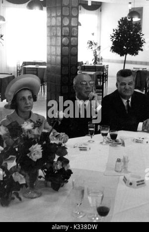 Während der Associazione Italo-Americana Mittagessen an der Schule der Erweiterte Internationale Studien (ORKB) in Washington, DC, Präsident der Johns Hopkins University Milton Stover Eisenhower - rund 62 Jahre des Alters - sitzt an einem Tisch, seine Hand halten zu seinem Kinn, mit seiner Frau zu seiner Rechten einen Hut tragen, und rauchen eine Zigarette, und ein anderer Gast zu seiner Linken und Weingläser um ein Blumenstrauß auf dem Tisch, Washington, DC, 1965. Stockfoto