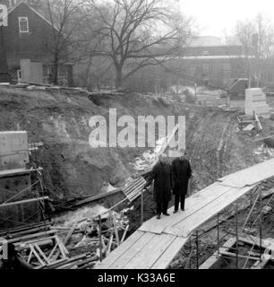 In den frühen Phasen der Konstruktion der Milton S Eisenhower Bibliothek an der Johns Hopkins University, zwei Mitarbeiter der Universität tragen wolle Mäntel stehen auf einem Laufsteg von Brettern über Gerüste, Überprüfung der Fortschritte des Projekts, mit Stapeln von Balken und anderen Materialien, um das Innere der Schmutz Grube liegen, die die Grundlage des Gebäudes, mit dem Merrick Scheune, ein Spielhaus auf dem Campus, im Hintergrund auf der linken Seite, und Whitehead Hall, ein Labor und Wissenschaft Gebäude, rechts, Baltimore, Maryland, 1964. Stockfoto