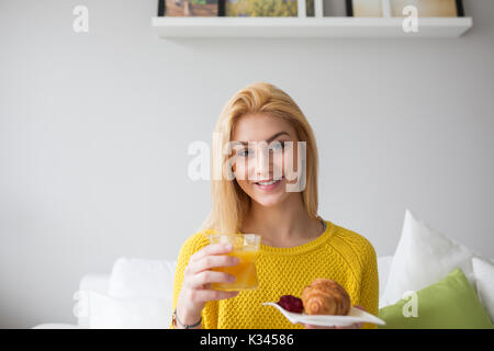 Ein Foto der jungen Frau im gelben Pullover Holding eine Platte mit Croissant und Marmelade und ein Glas Saft. Sie lächelt. Stockfoto