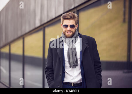 Ein Foto der Jungen, modischen buinessman Neben einige Gebäude mit seinen Händen in den Taschen und lächelnd. Stockfoto