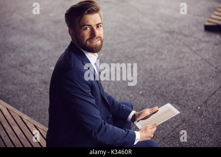 Ein Foto der junge Geschäftsmann im Anzug auf der Bank sitzen und auf Tablet. Er lächelt. Stockfoto