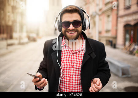 Ein Foto der jungen, trendigen Mann stand auf dem Marktplatz und die Musik durch weiße Ohrhörer. Er ist glücklich. Stockfoto