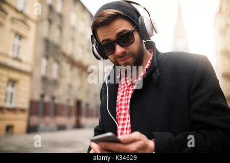 Ein Foto der jungen, trendigen Mann Hören von Musik über Kopfhörer und über Handy. Stockfoto