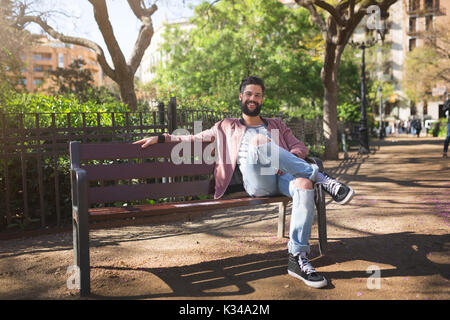 Ein Foto der junge Mann sitzt auf der Bank im Park. Er lächelt. Stockfoto