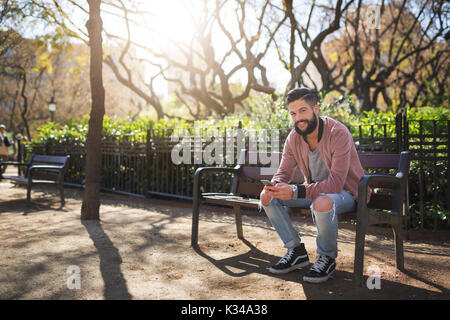 Ein Foto der junge Mann sitzt auf der Bank im Park und texting und seine Freunde. Er lächelt. Stockfoto