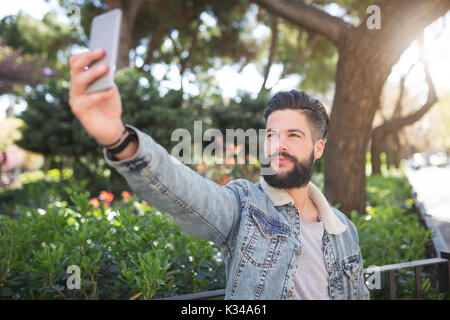 Ein Foto der jungen, gutaussehenden Mann stand auf der Allee im Park und nehmen sich selbst ein Foto von seinem Handy. Stockfoto