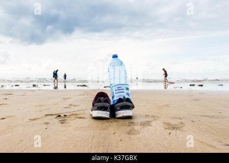 Low Angle Nahaufnahme einer Wasserflasche und Schuhe von einem Surfer auf einem sandigen Strand bei Ebbe links, Westward Ho!, Devon, England, Großbritannien Stockfoto