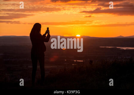 Eine Frau nimmt ein Foto auf den Sonnenuntergang mit Ihrem Smart Phone aus Coaley Peak in Gloucestershire suchen in Richtung des Flusses Severn und Wald von Dean. Stockfoto