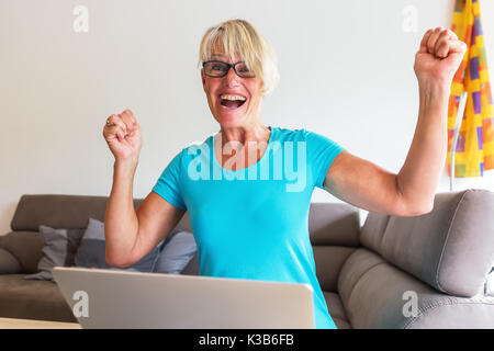 Bild von einer reifen Frau sitzt, die Freude mit erhobenen Händen vor einem Laptop ist Stockfoto