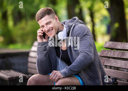 Ein Foto von einem jungen Mann in grauen Sweatshirt auf der Bank sitzen und sprechen auf das Handy. Stockfoto