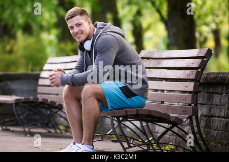 Ein Foto der jungen, gutaussehenden Mann sitzt auf der Bank im Park und entspannend. Stockfoto