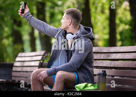 Ein Foto der junge Mann sitzt auf der Bank im Park und einen selfie. Stockfoto