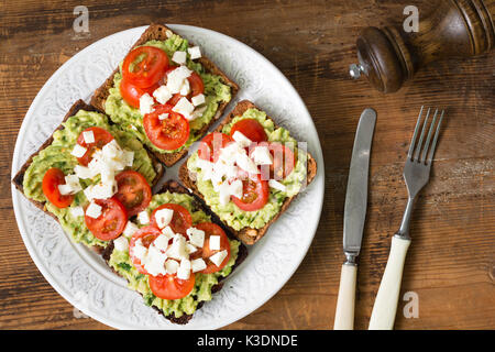 Toast mit Guacamole, Tomaten und zerkrümeltem Fetakäse auf weißem Schild auf hölzernen Tisch. nach oben anzeigen. gesunde Ernährung, gesunder Snack Konzept Stockfoto