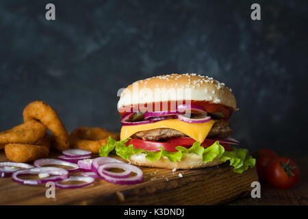 Cheeseburger und Zwiebelringe auf Holz Schneidebrett über dunklen Hintergrund. Detailansicht, selektive konzentrieren. Fast food Konzept Stockfoto