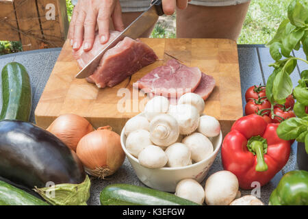 Vorderansicht des Frau schneiden Fleisch auf einem Holzbrett. Verschiedene Arten von Gemüse liegen auf dem Tisch. Stockfoto
