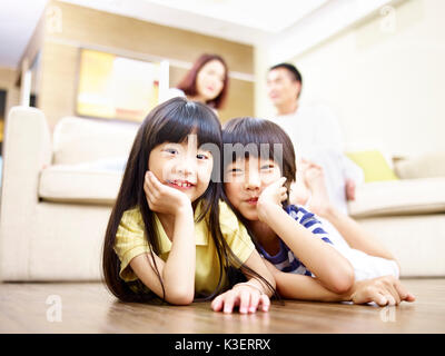 Asiatische Bruder und Schwester liegen auf der Vorderseite auf den Boden mit den Eltern im Hintergrund. Stockfoto