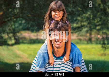 Vater trägt Lachen Tochter mit braunen langen Haaren auf den Schultern Stockfoto