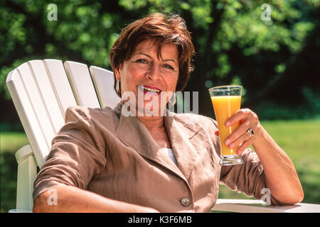 Frau im mittleren Alter sitzt im Garten Stuhl mit einem Glas Orangensaft Stockfoto