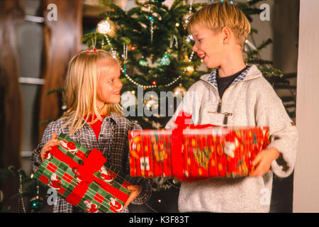 Zwei Kinder vor einem Weihnachtsbaum mit Geschenken in der Hand Stockfoto