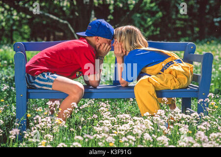 Junge und Mädchen sitzen in einem Garten auf der Armlehne eines Garten Bank und Flüstern Stockfoto