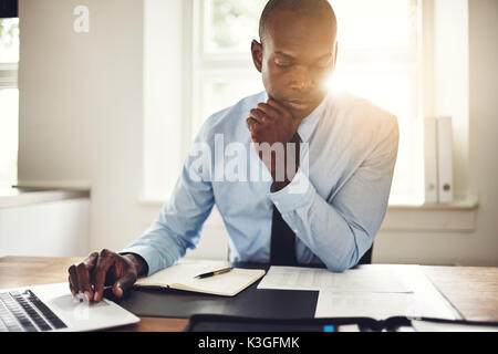 Junge afrikanische Executive arbeitet an einem Laptop und Lesen von Dokumenten, während an seinem Schreibtisch in einem Büro sitzen Stockfoto